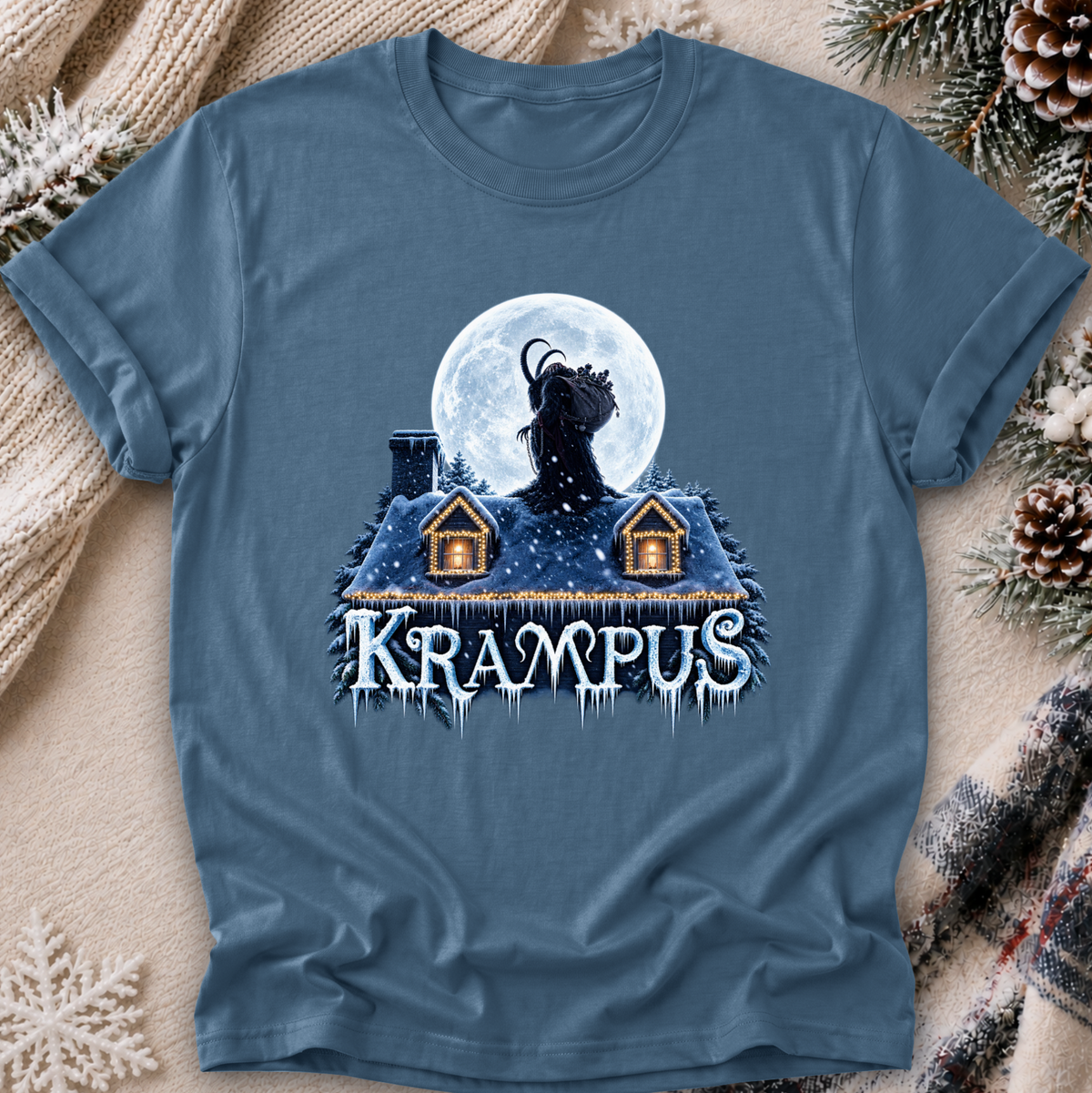 A slate blue graphic t-shirt laid flat on a cozy holiday background with a knitted blanket, pinecones, and evergreen branches. The graphic features a goat-headed monster (Krampus) howling at a full moon above a snowy house with the word 'Krampus' written below in icy white letters.