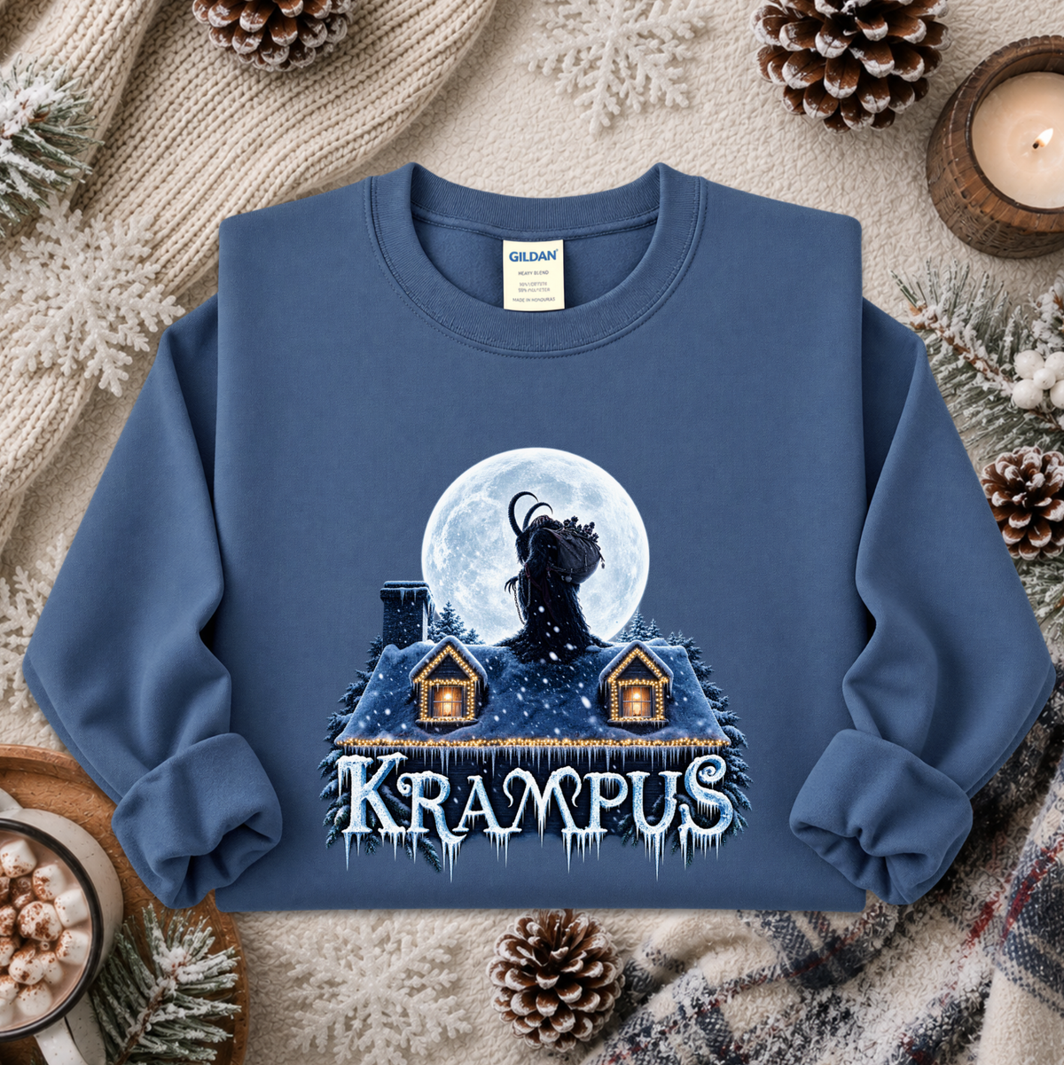 A blue crewneck sweatshirt featuring a graphic of a black goat under a full moon above a snowy house, with the word 'KRAMPUS' in icy letters. The sweatshirt is displayed in a cozy flat-lay setting with pinecones, a candle, and a knit blanket.