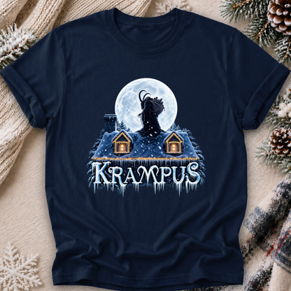 A navy blue graphic t-shirt laid flat on a cozy holiday-themed background of knitted blankets, pinecones, and evergreen branches. The graphic features a silhouette of a goat-like creature howling at a full moon above two small houses, with the word 'KRAMPUS' written below in icy white letters with blue icicle details.