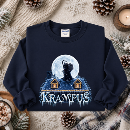A navy blue crewneck sweatshirt with a graphic of a goat-like creature in front of a full moon above two small houses, with the word 'KRAAMPUS' in icy letters below. The sweatshirt is displayed in a flat-lay arrangement with a cozy winter background including pinecones, a candle, and a knit blanket.
