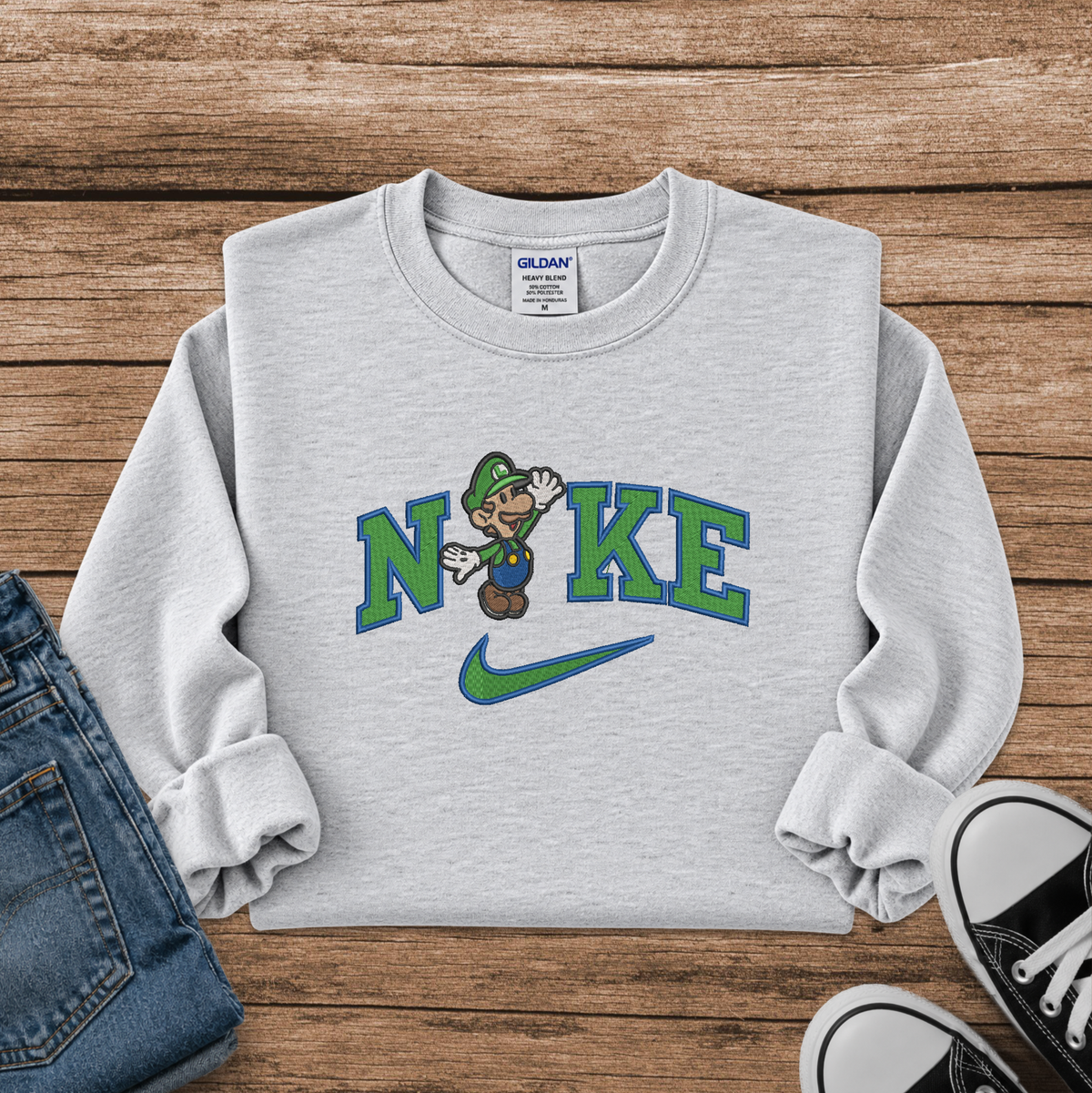 A light heather gray crewneck sweatshirt laid flat on a rustic wooden background, featuring a 'N' and 'K' styled with a green and blue Nike swoosh and a cartoon Luigi character. The sweatshirt is styled with blue jeans and black sneakers.
