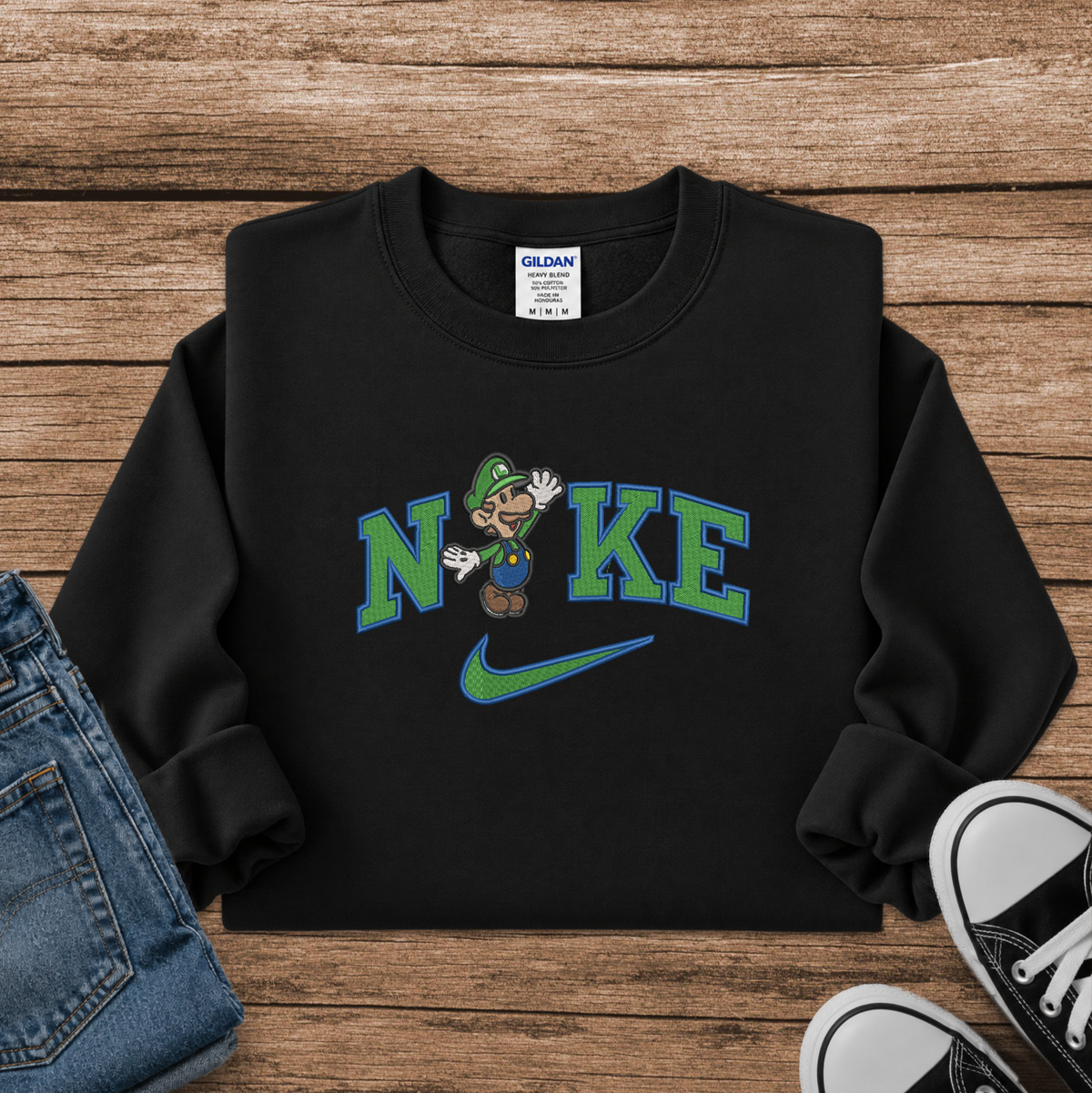 A black crewneck sweatshirt featuring a 'Nike' parody graphic with Luigi instead of the Nike swoosh and a cartoon character, styled in a flat lay on a wooden background with jeans and sneakers.