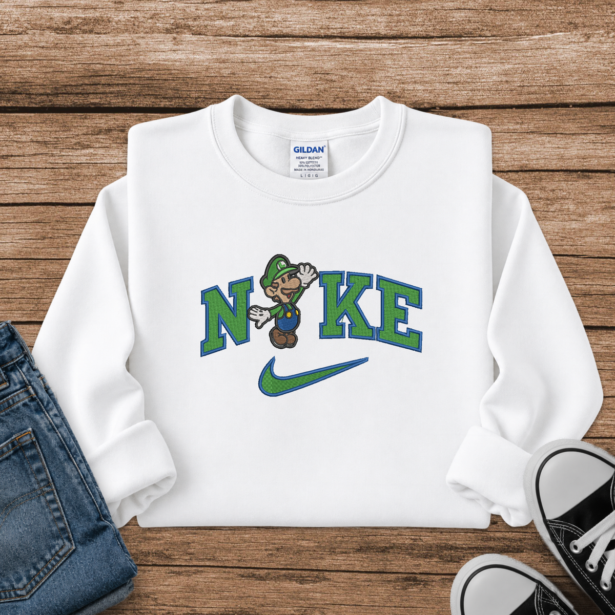 A white long-sleeve crewneck sweatshirt laid flat on a rustic wooden surface, styled with blue jeans and black sneakers. The sweatshirt features a graphic print of the word 'NIKE' in green and blue, with a cartoon Luigi replacing the letter 'I', and a green Nike swoosh underneath.