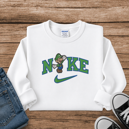 A white long-sleeve crewneck sweatshirt laid flat on a rustic wooden surface, styled with blue jeans and black sneakers. The sweatshirt features a graphic print of the word 'NIKE' in green and blue, with a cartoon Luigi replacing the letter 'I', and a green Nike swoosh underneath.