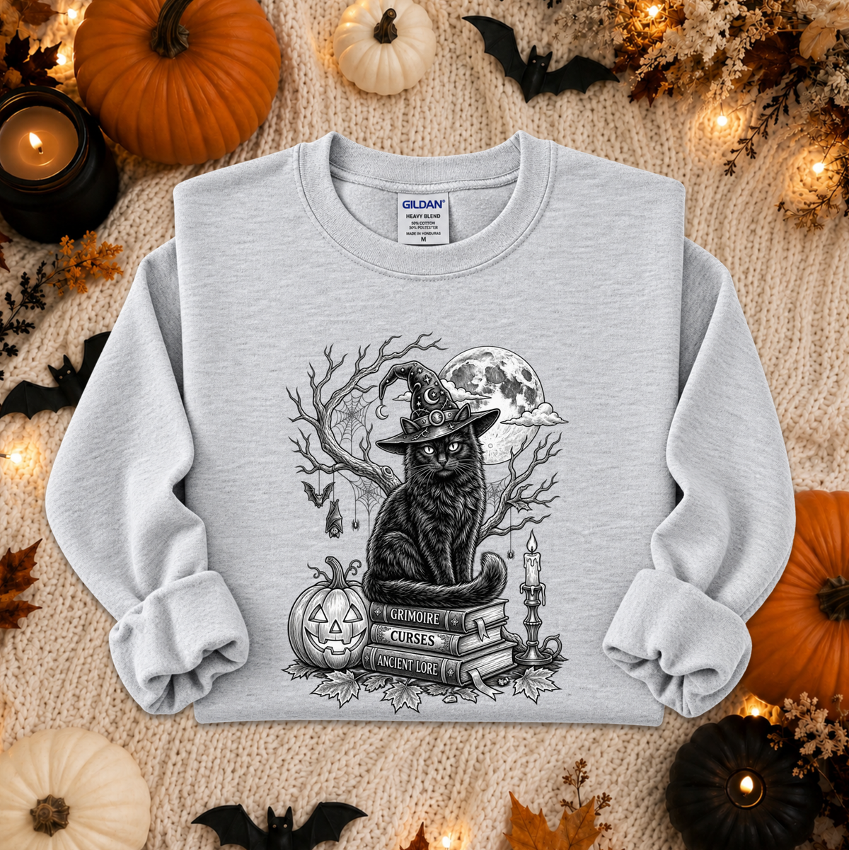 A light gray crewneck sweatshirt with a black and white graphic of a witch cat sitting on books, surrounded by Halloween decorations like pumpkins, candles, and bats, laid flat on a knitted blanket.