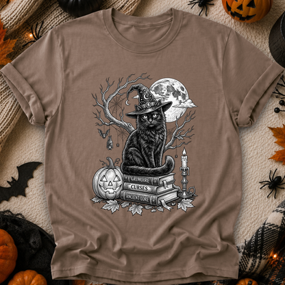 A taupe brown short-sleeve crewneck t-shirt featuring a detailed black and white graphic of a witch cat sitting on books next to a jack-o'-lantern and a full moon. The shirt is displayed in a flat-lay Halloween setting with props like a knit blanket, plastic spiders, and pumpkins.