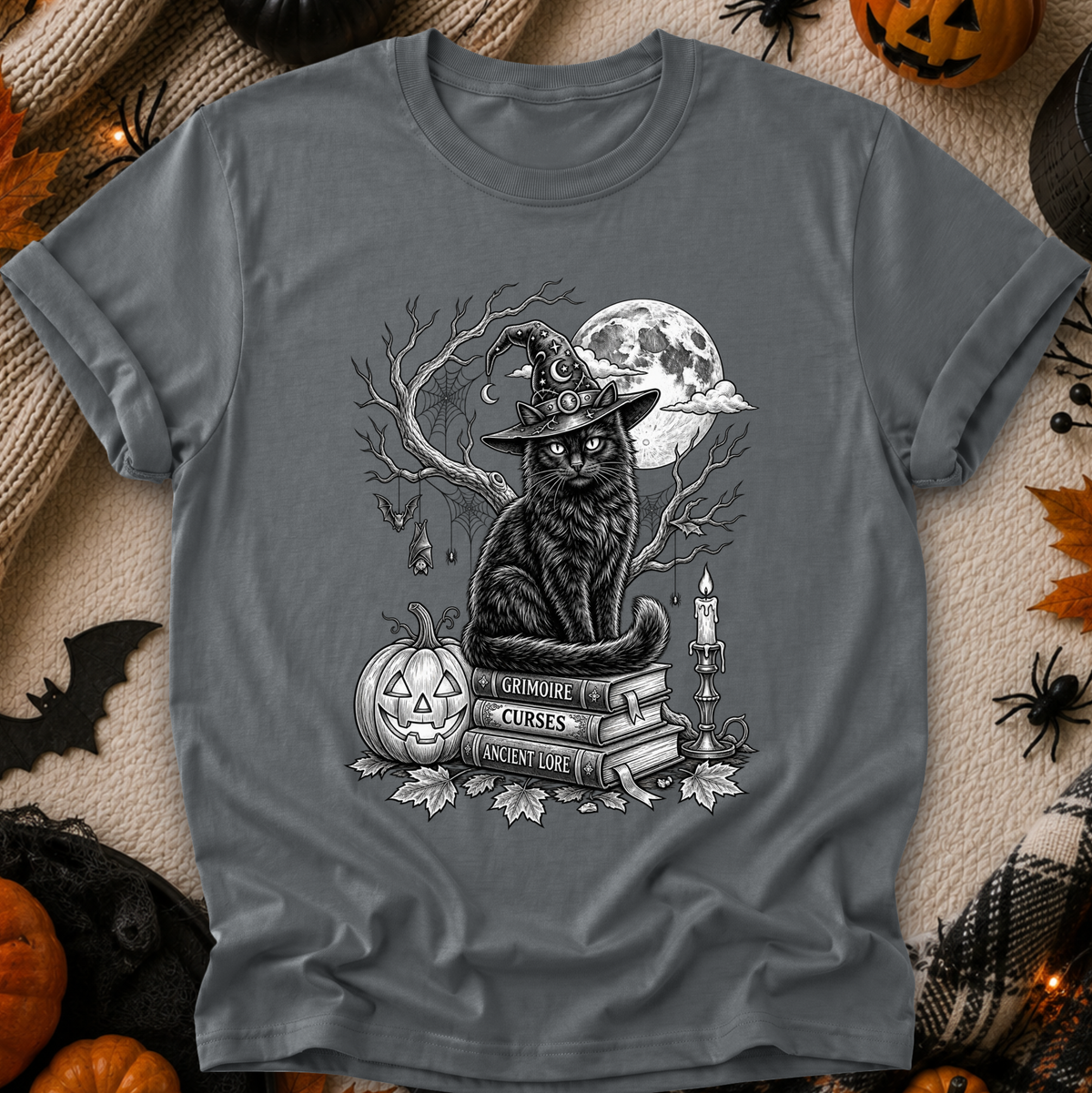A dark gray graphic t-shirt featuring a black cat dressed as a witch sitting on books next to a jack-o'-lantern and a full moon. The t-shirt is displayed in a flat-lay Halloween setting with pumpkins, spider decorations, and a beige knit blanket.
