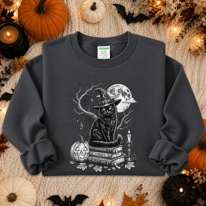 A dark charcoal gray crewneck sweatshirt featuring a detailed black and white graphic of a witch cat sitting on books with a pumpkin and full moon. The sweatshirt is styled in a cozy autumn flat-lay setting with pumpkins, candles, and leaves on a textured blanket.