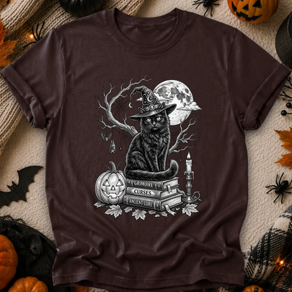 A dark brown short-sleeve t-shirt with a detailed black and white graphic of a witch cat sitting on books next to a jack-o'-lantern and full moon. The shirt is displayed in a flat-lay Halloween setting with pumpkins, spiderwebs, and a beige blanket.