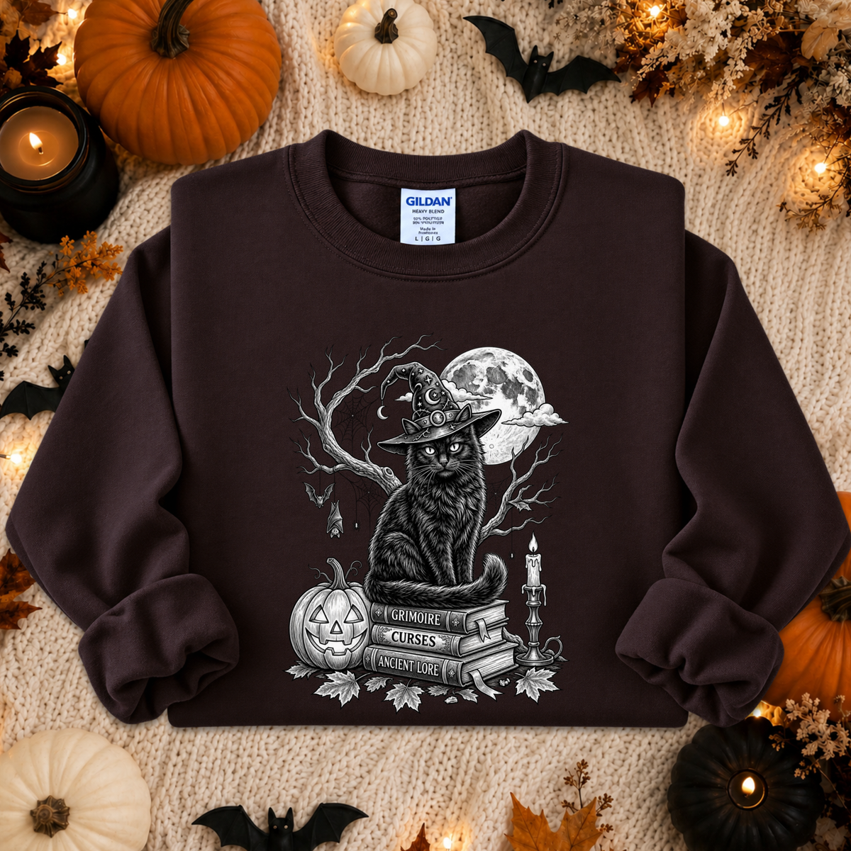 A dark brown Gildan sweatshirt with a Halloween-themed graphic print of a witch cat sitting on books, displayed on a cozy autumn-themed background with pumpkins, candles, and bats.