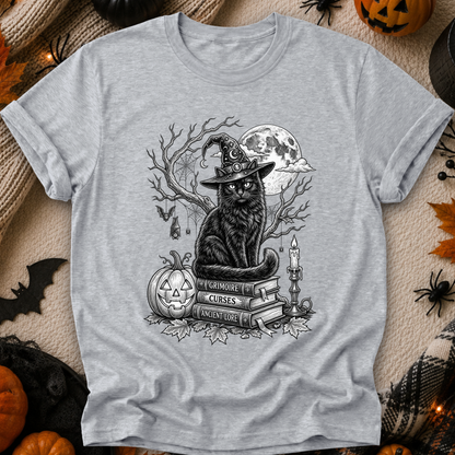 A light heather gray short-sleeve T-shirt laid flat with a Halloween-themed black line-art graphic on the chest showing a witch cat, moon, pumpkin, books, and candles. The shirt is photographed on a cozy beige fabric background with autumn props like pumpkins, spiderwebs, and leaves.
