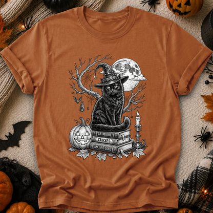 An orange crewneck t-shirt with a black and white graphic of a witch cat sitting on books next to a jack-o'-lantern and full moon. The shirt is displayed in a flat-lay Halloween setting with pumpkins, spiderwebs, and a cozy blanket.