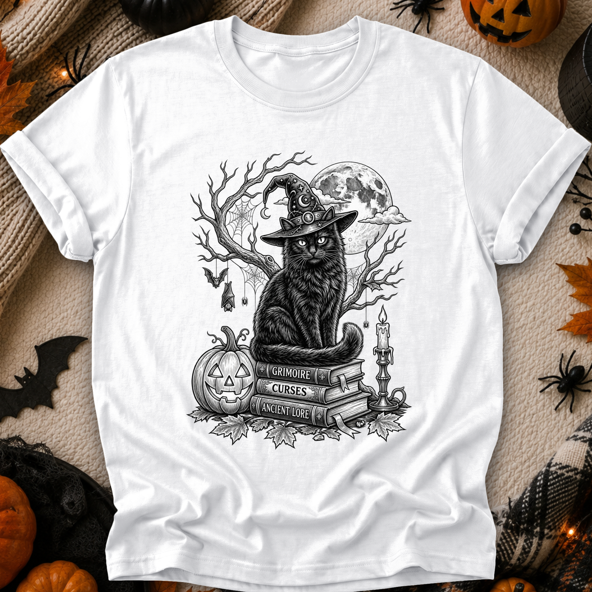 A white short-sleeve crewneck T-shirt laid flat, featuring a black-and-gray graphic of a witch cat sitting on books beside a jack-o'-lantern, with a full moon and bare branches in the background. The shirt is styled on a beige fabric surface with Halloween props including pumpkins, spiders, and a black spider decoration around the edges.