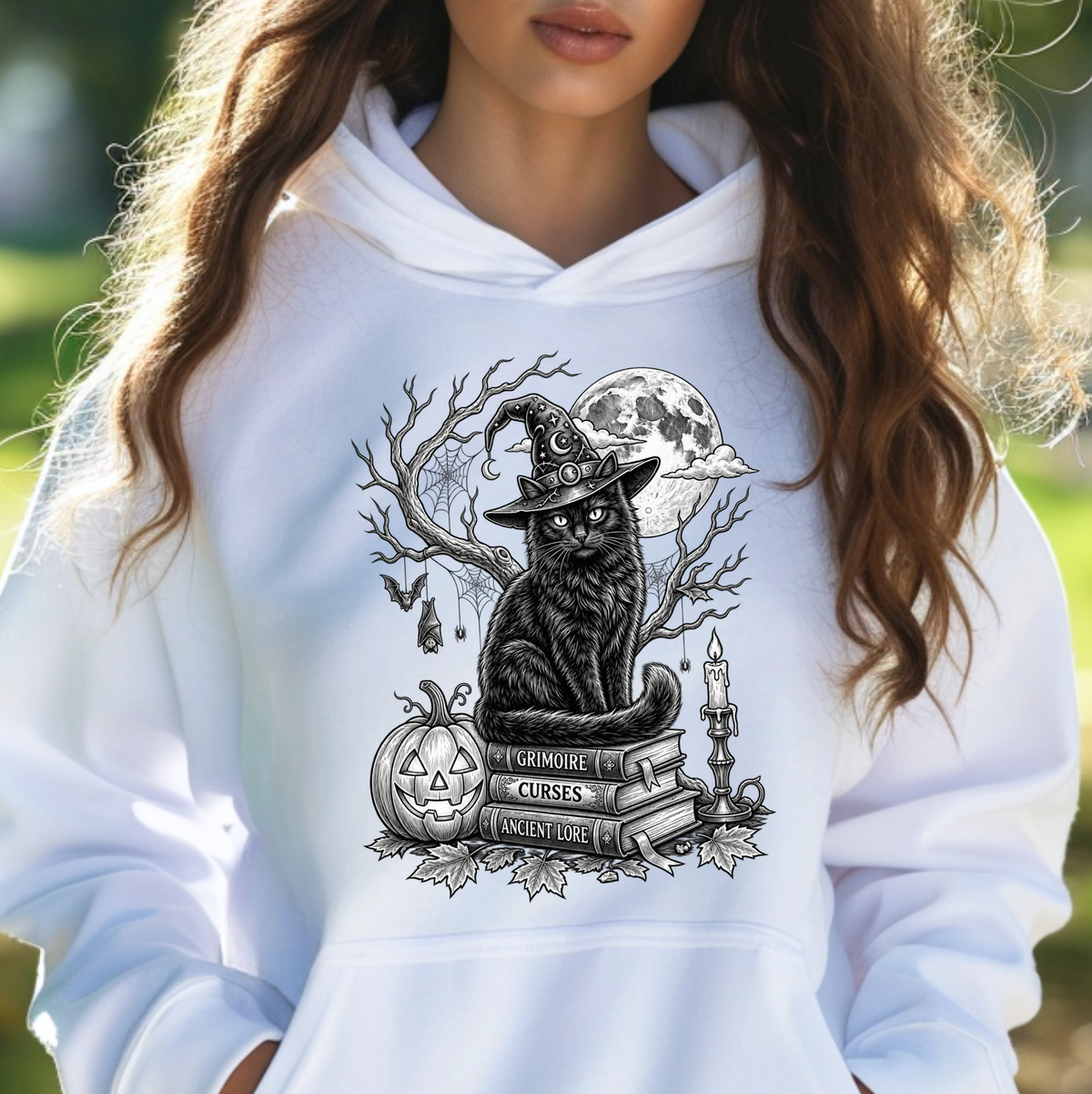 A woman wearing a white hoodie with a black and gray graphic of a witch cat sitting on books with a pumpkin and full moon.