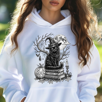 A woman wearing a white hoodie with a black and gray graphic of a witch cat sitting on books with a pumpkin and full moon.