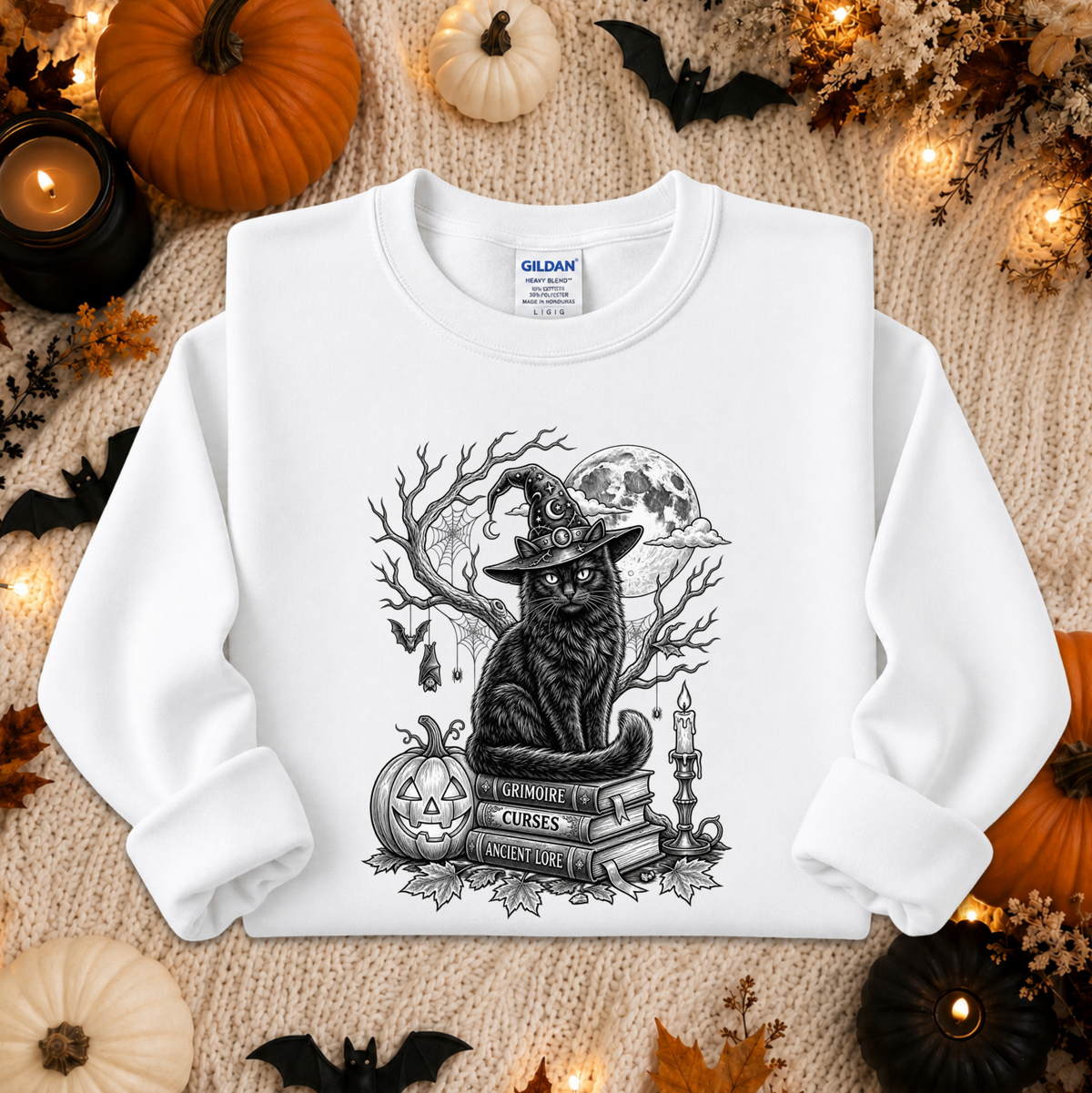 A white long-sleeve sweatshirt laid flat on a beige knit surface, surrounded by Halloween props including pumpkins, candles, leaves, and bats. The sweatshirt has a large black graphic of a witch cat sitting on books with a pumpkin and moon in the background.