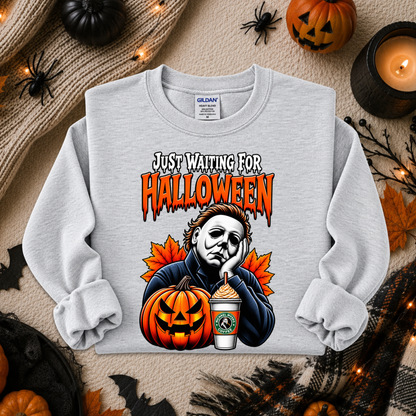 A light gray crewneck sweatshirt with a graphic print of Michael Myers sitting behind a jack-o'-lantern and a Starbucks cup, with the text 'JUST WAITING FOR HALLOWEEN'. The sweatshirt is displayed in a flat-lay arrangement with Halloween props including pumpkins, a candle, and spider decorations on a beige knit blanket.