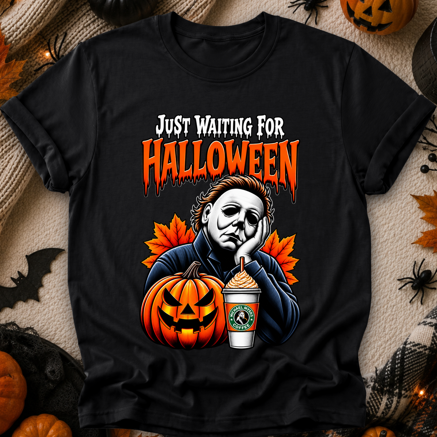 A black short-sleeve graphic T-shirt laid flat on a textured beige fabric with Halloween props around it. The shirt features a colorful print of Michael Myers looking bored beside a jack-o'-lantern and a Starbucks cup, with the text 'JUST WAITING FOR HALLOWEEN' in white and orange.