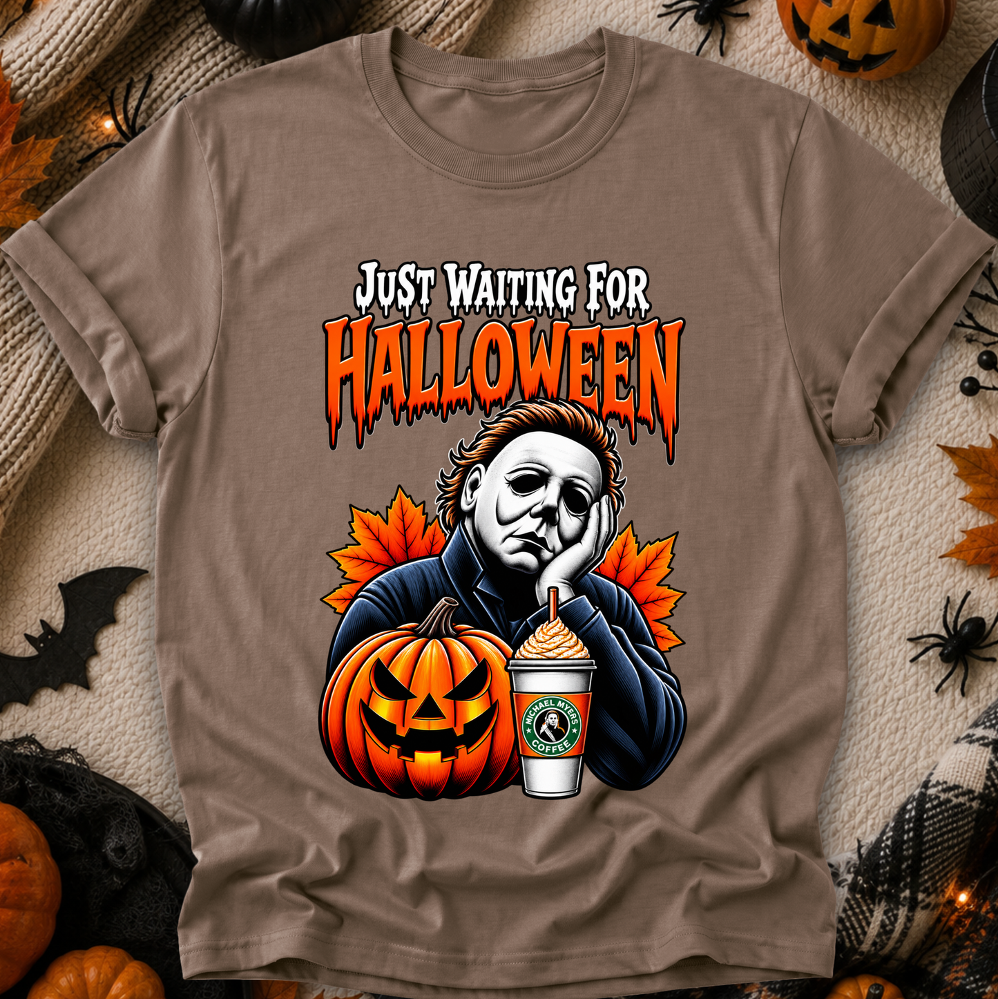 A taupe brown graphic t-shirt featuring a Halloween-themed illustration of Michael Myers, a jack-o'-lantern, a Starbucks cup, and orange maple leaves, with the text 'JUST WAITING FOR HALLOWEEN'. The t-shirt is displayed in a flat-lay arrangement surrounded by seasonal props like pumpkins, a spider, and a black cat.