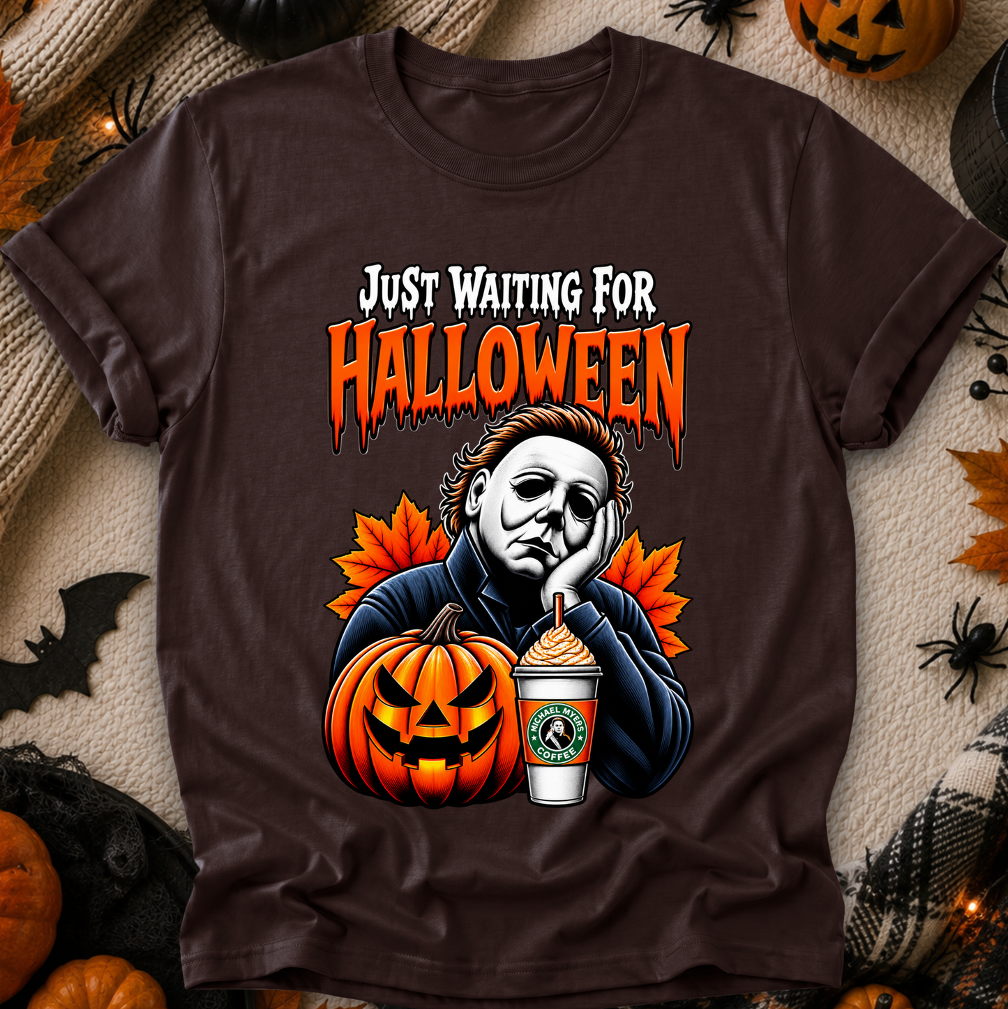 A dark brown graphic t-shirt featuring a Halloween-themed design with the text 'JUST WAITING FOR HALLOWEEN' and an illustration of Michael Myers, a jack-o'-lantern, and a Starbucks cup. The shirt is displayed in a flat-lay arrangement with seasonal props like pumpkins, a spider, and a bat.