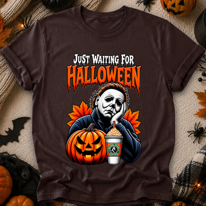 A dark brown graphic t-shirt featuring a Halloween-themed design with the text 'JUST WAITING FOR HALLOWEEN' and an illustration of Michael Myers, a jack-o'-lantern, and a Starbucks cup. The shirt is displayed in a flat-lay arrangement with seasonal props like pumpkins, a spider, and a bat.