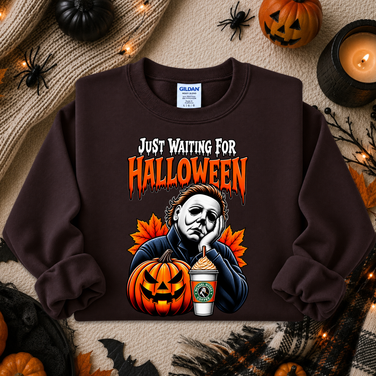 A dark brown Gildan sweatshirt featuring a Halloween graphic of Michael Myers looking bored next to a jack-o'-lantern and a pumpkin spice latte, with the text 'JUST WAITING FOR HALLOWEEN' in orange. The sweatshirt is displayed in a flat-lay Halloween setting with pumpkins, a candle, and spider decorations.