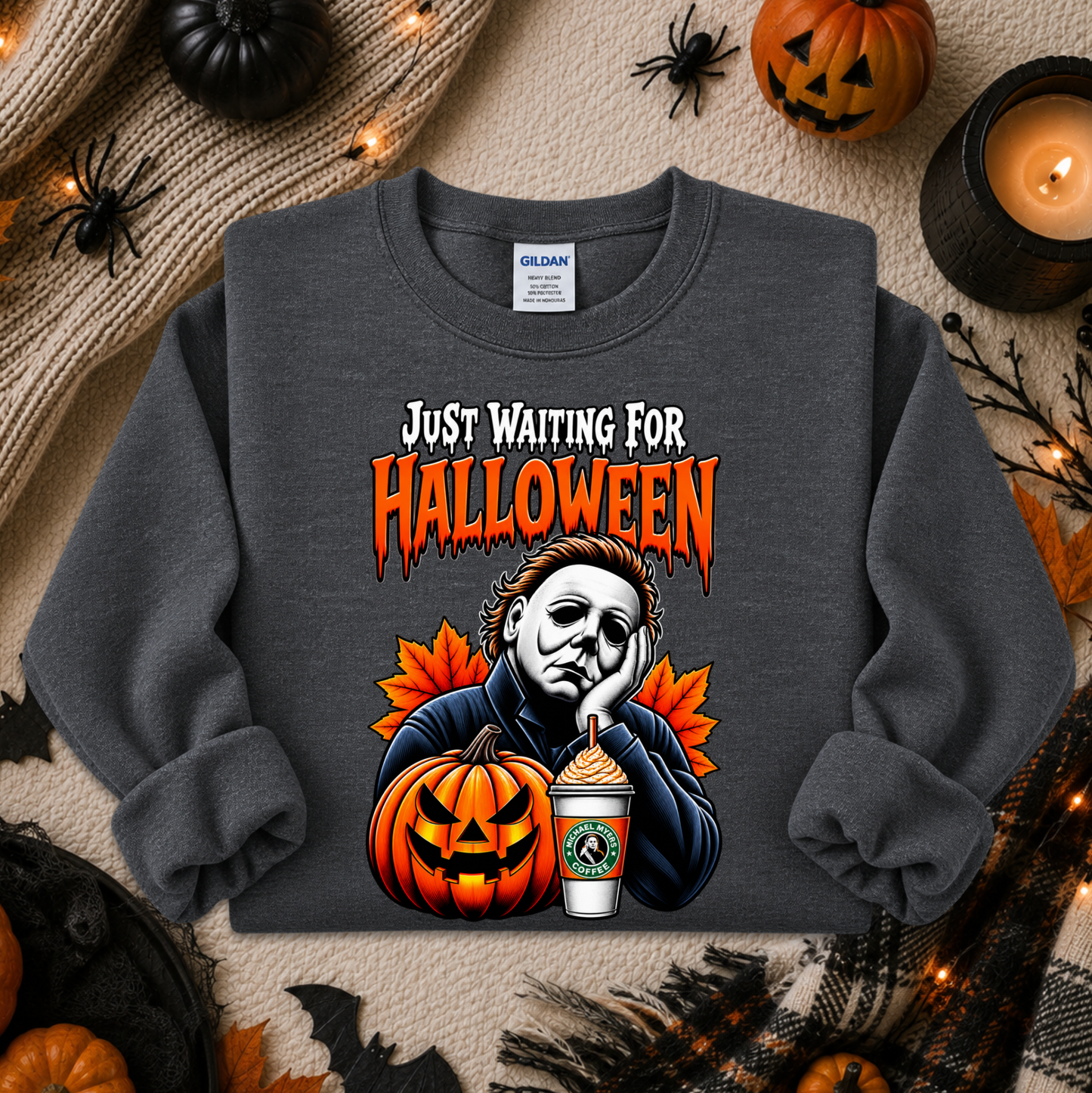A dark gray sweatshirt with a Halloween graphic print featuring Michael Myers, a jack-o'-lantern, and a coffee cup, styled in a flat lay with autumn props.