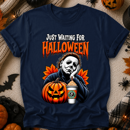 A navy blue graphic t-shirt featuring a Halloween-themed design with the text 'JUST WAITING FOR HALLOWEEN' in orange and white, an illustration of Michael Myers, a jack-o'-lantern, and a Starbucks cup, displayed on a cozy beige knit blanket surrounded by Halloween decorations.