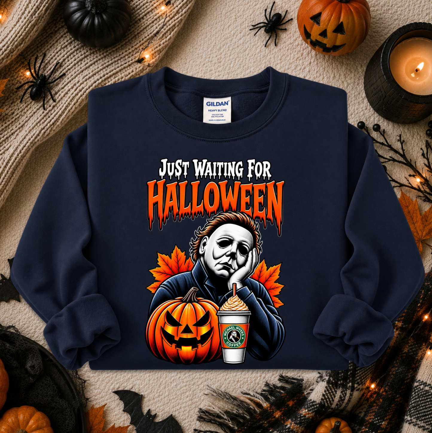 A navy blue crewneck sweatshirt featuring a Michael Myers graphic with the text 'JUST WAITING FOR HALLOWEEN' in orange. The sweatshirt is styled in a flat lay with Halloween props including pumpkins, a candle, and autumn leaves on a beige knit blanket.