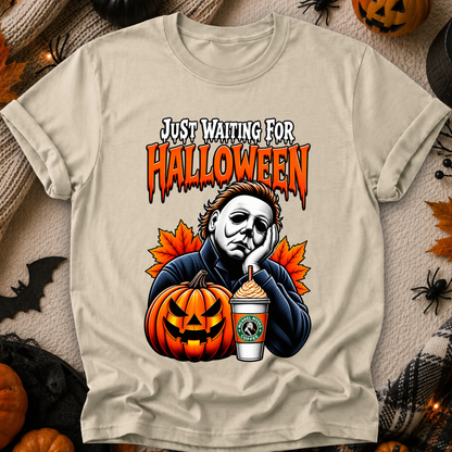 A beige graphic t-shirt featuring a Halloween-themed design with the text 'JUST WAITING FOR HALLOWEEN' above an illustration of Michael Myers, a jack-o'-lantern, and a Starbucks drink, displayed on a cozy autumn background with pumpkins and spider decorations.