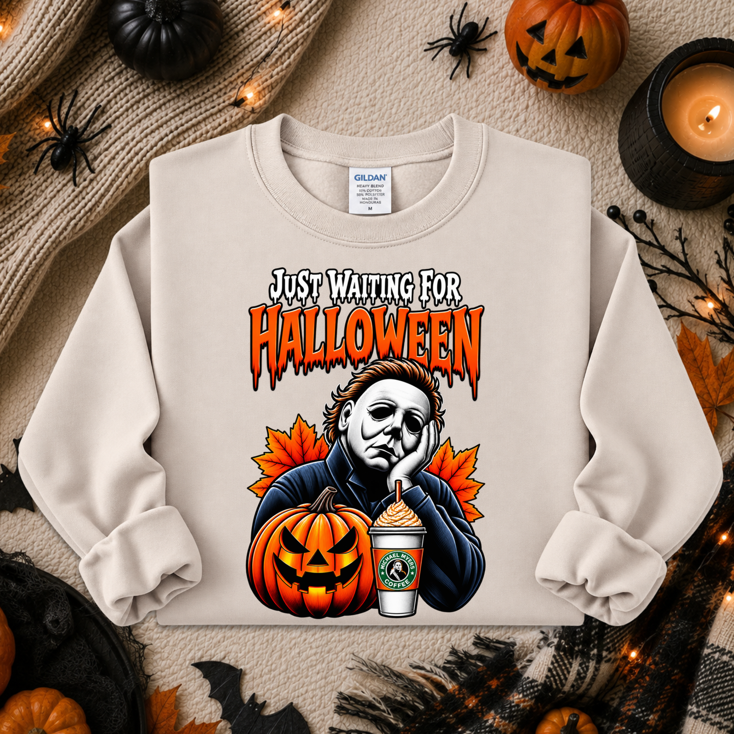 A cream-colored crewneck sweatshirt featuring a graphic of Michael Myers looking sad and bored with the text 'JUST WAITING FOR HALLOWEEN' above him. The sweatshirt is displayed in a flat-lay Halloween setting with pumpkins, a candle, and a knit sweater.
