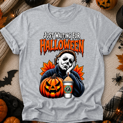 A heather gray graphic t-shirt featuring a Halloween-themed design with the text 'JUST WAITING FOR HALLOWEEN' in orange dripping letters, an illustration of Michael Myers, a jack-o'-lantern, and a Starbucks-style iced coffee, presented in a flat-lay with seasonal props.