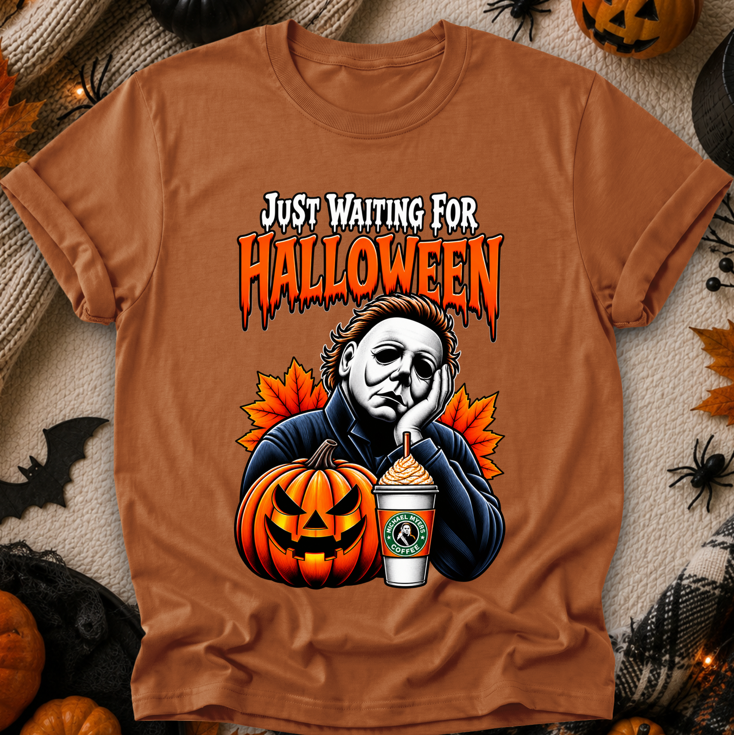 A brown graphic t-shirt featuring a Halloween-themed illustration of Michael Myers, a jack-o'-lantern, a Starbucks cup, and orange maple leaves, with the text 'JUST WAITING FOR HALLOWEEN'. The shirt is displayed in a flat-lay arrangement on a textured beige blanket surrounded by seasonal props like pumpkins, a spider, and a black spiderweb.
