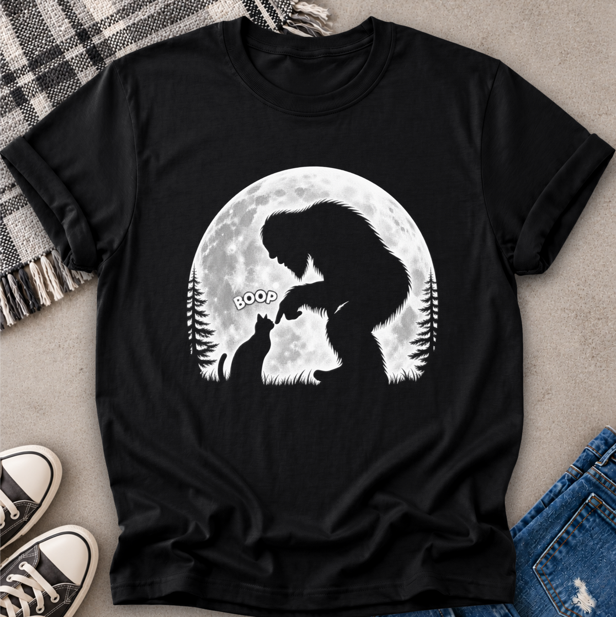 A black short-sleeve graphic T-shirt laid flat on a light gray surface with a plaid blanket, sneakers, and blue jeans around it. The shirt features a large white circular moon graphic with a dark silhouette of a big cat and a small dog, plus the word 'BOOP' in a speech bubble.