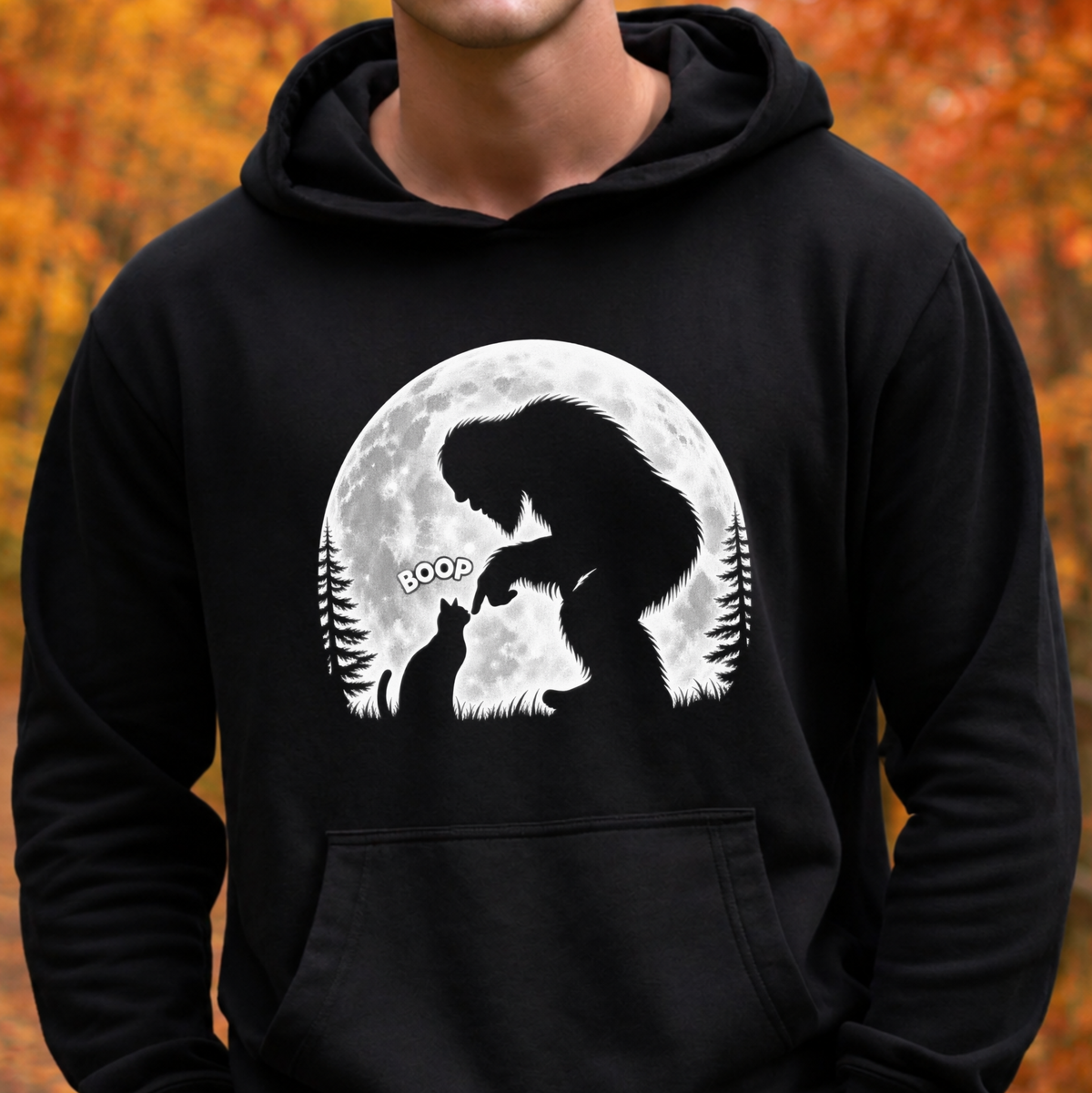 A person wearing a black pullover hoodie with a white graphic print of Bigfoot and a cat in front of a full moon, set against a blurred autumn foliage background.