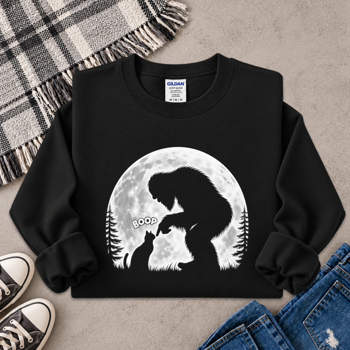 A black crewneck sweatshirt with a white graphic of Bigfoot and a cat in front of a full moon, styled in a flat lay with jeans, sneakers, and a plaid blanket on a gray concrete floor.