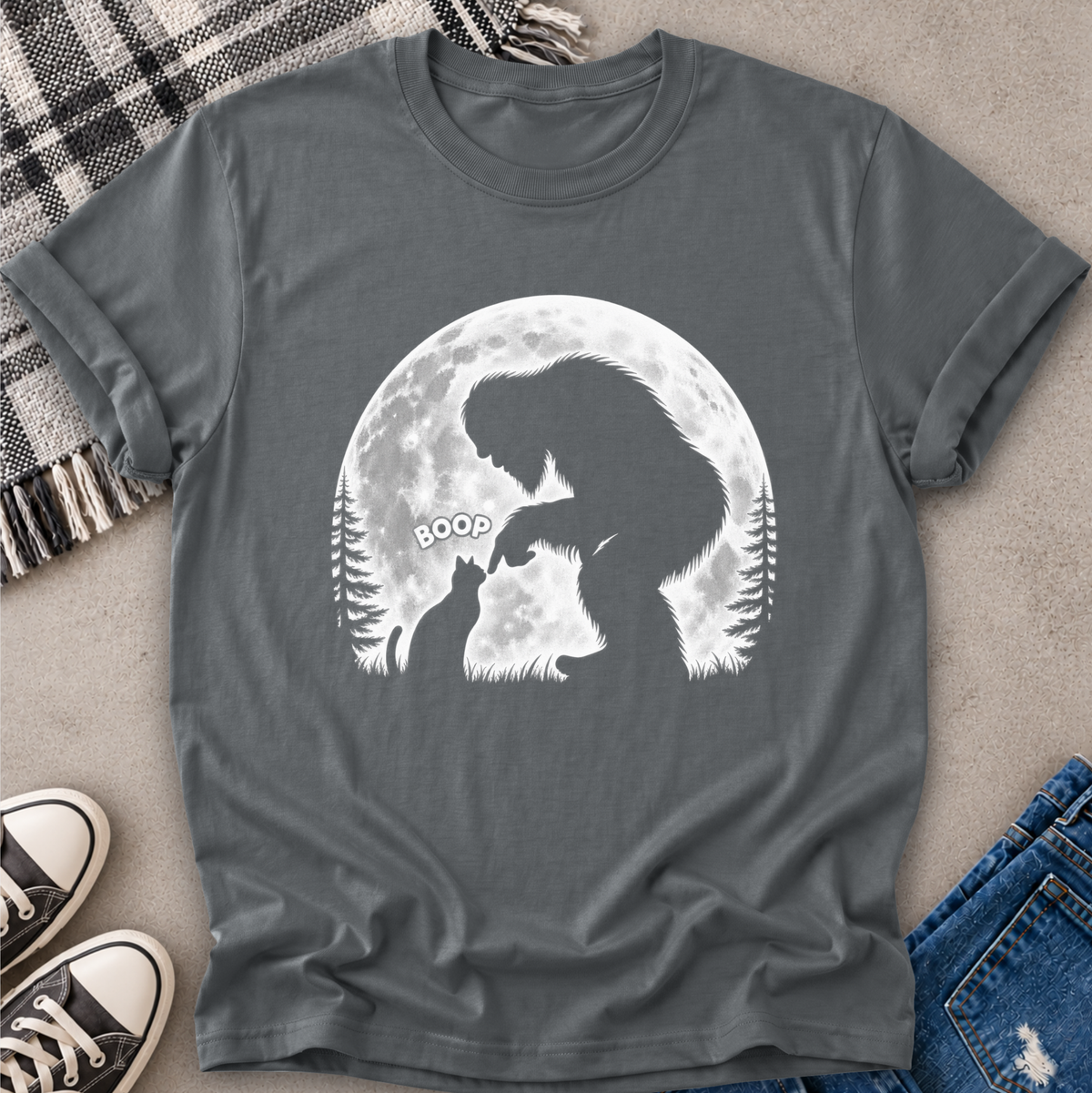 A dark gray crewneck t-shirt laid flat on a textured gray surface, featuring a white graphic of a Bigfoot silhouette interacting with a dog in front of a full moon, with 'BOOP' text. The shirt is styled with a plaid blanket, black sneakers, and blue jeans.