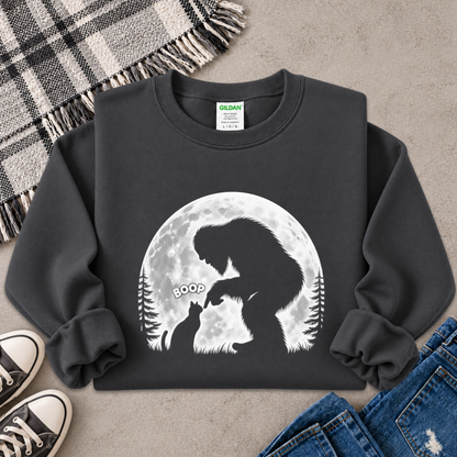 A dark charcoal gray crewneck sweatshirt with a graphic of a Bigfoot silhouette facing a cat in front of a full moon, styled in a flat lay with jeans, sneakers, and a plaid blanket on a concrete floor.