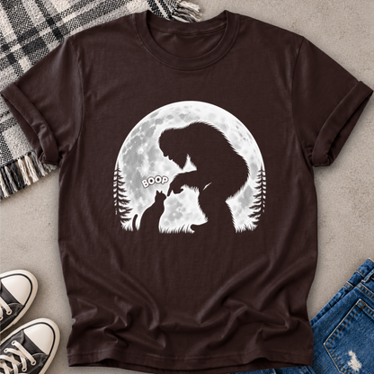 A navy blue short-sleeve t-shirt with a white graphic of a Bigfoot and a cat silhouette in front of a full moon, styled in a flat-lay with jeans, sneakers, and a plaid blanket.