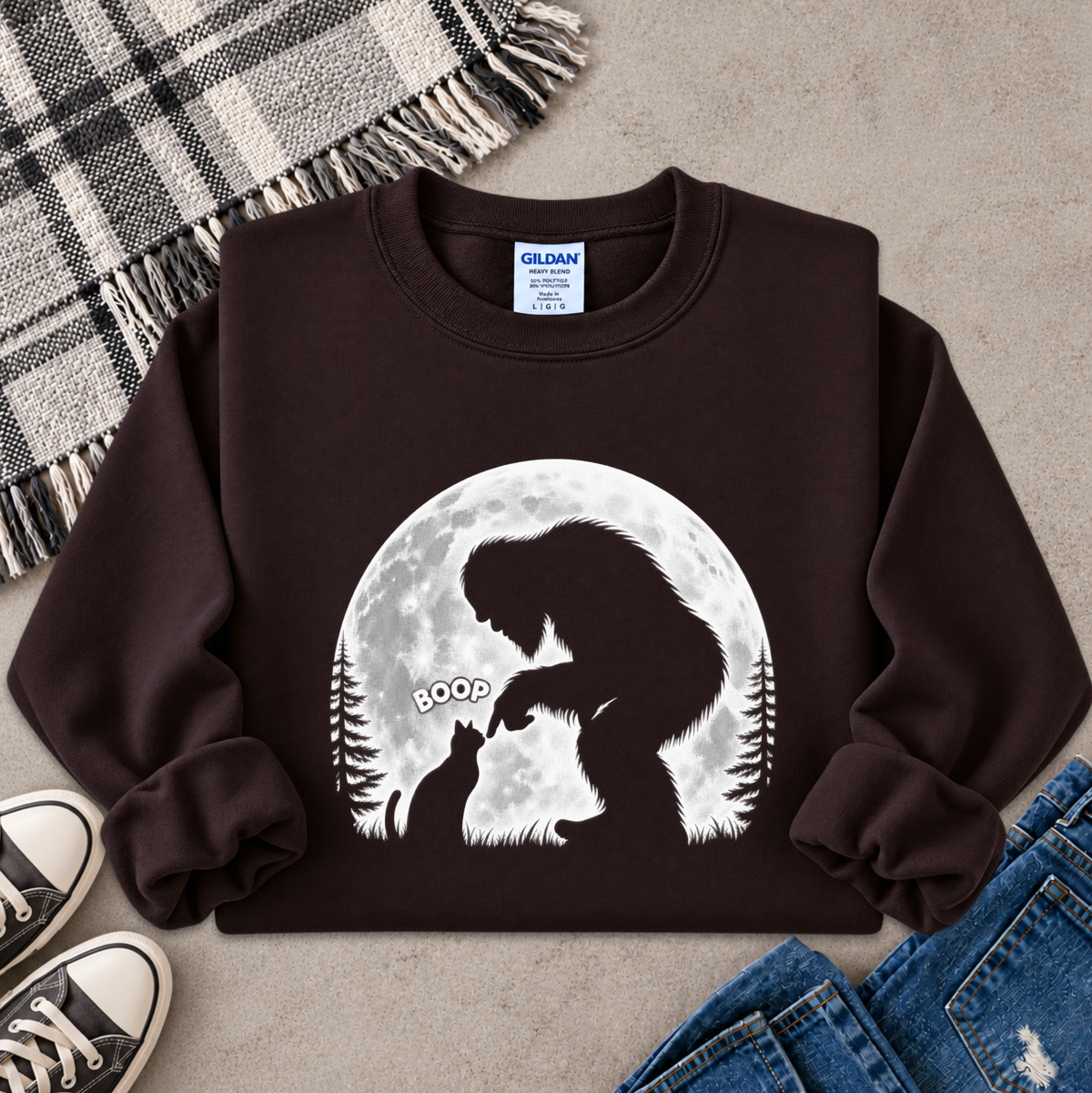 A dark brown crewneck sweatshirt featuring a white graphic of a Bigfoot silhouette looking at a cat in front of a full moon, styled in a flat lay with jeans, sneakers, and a plaid blanket on a gray concrete background.