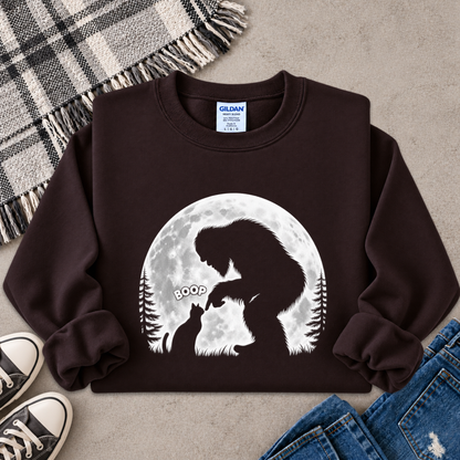 A dark brown crewneck sweatshirt featuring a white graphic of a Bigfoot silhouette looking at a cat in front of a full moon, styled in a flat lay with jeans, sneakers, and a plaid blanket on a gray concrete background.
