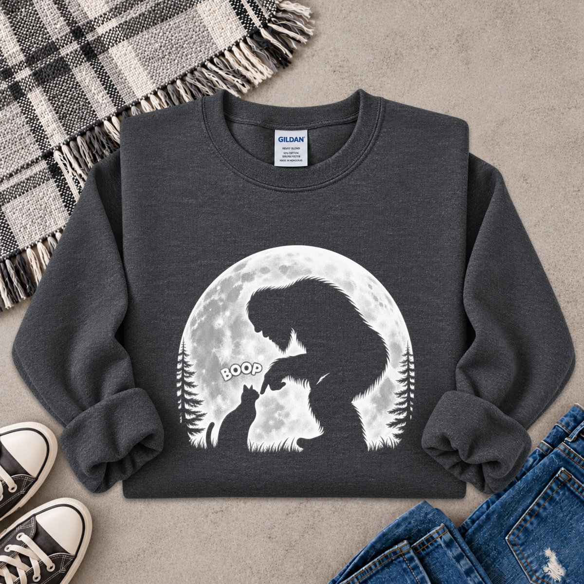 A dark charcoal gray sweatshirt with a large white graphic of a Bigfoot silhouette against a full moon, styled in a flat lay with jeans, sneakers, and a plaid blanket on a concrete floor.