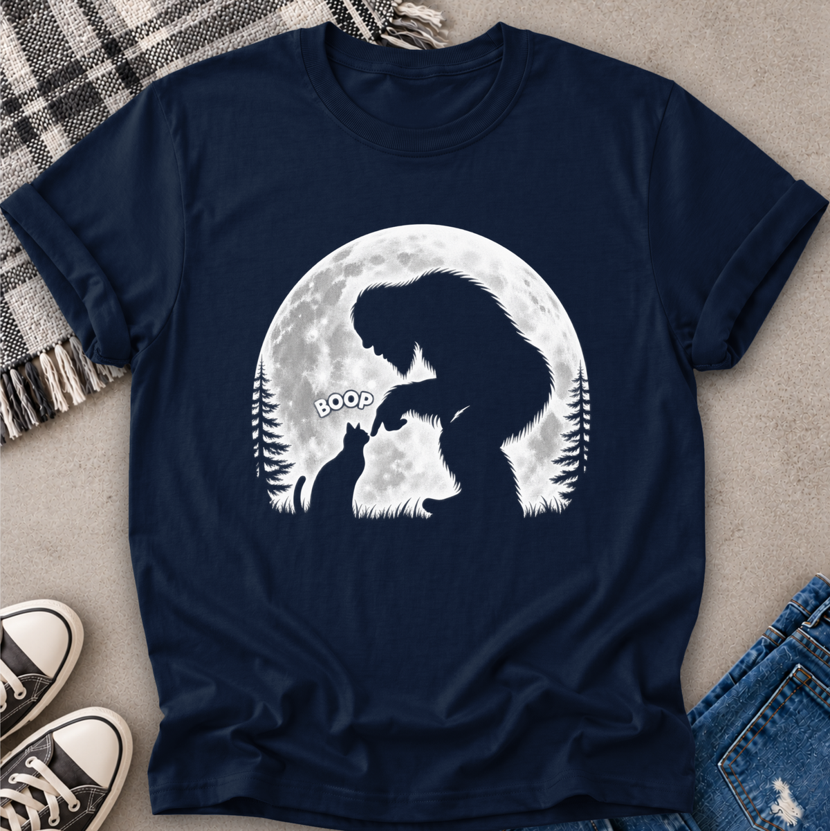 A navy blue short-sleeve t-shirt with a white graphic of a Bigfoot and a cat silhouette in front of a full moon, styled in a flat-lay with jeans, sneakers, and a plaid blanket.