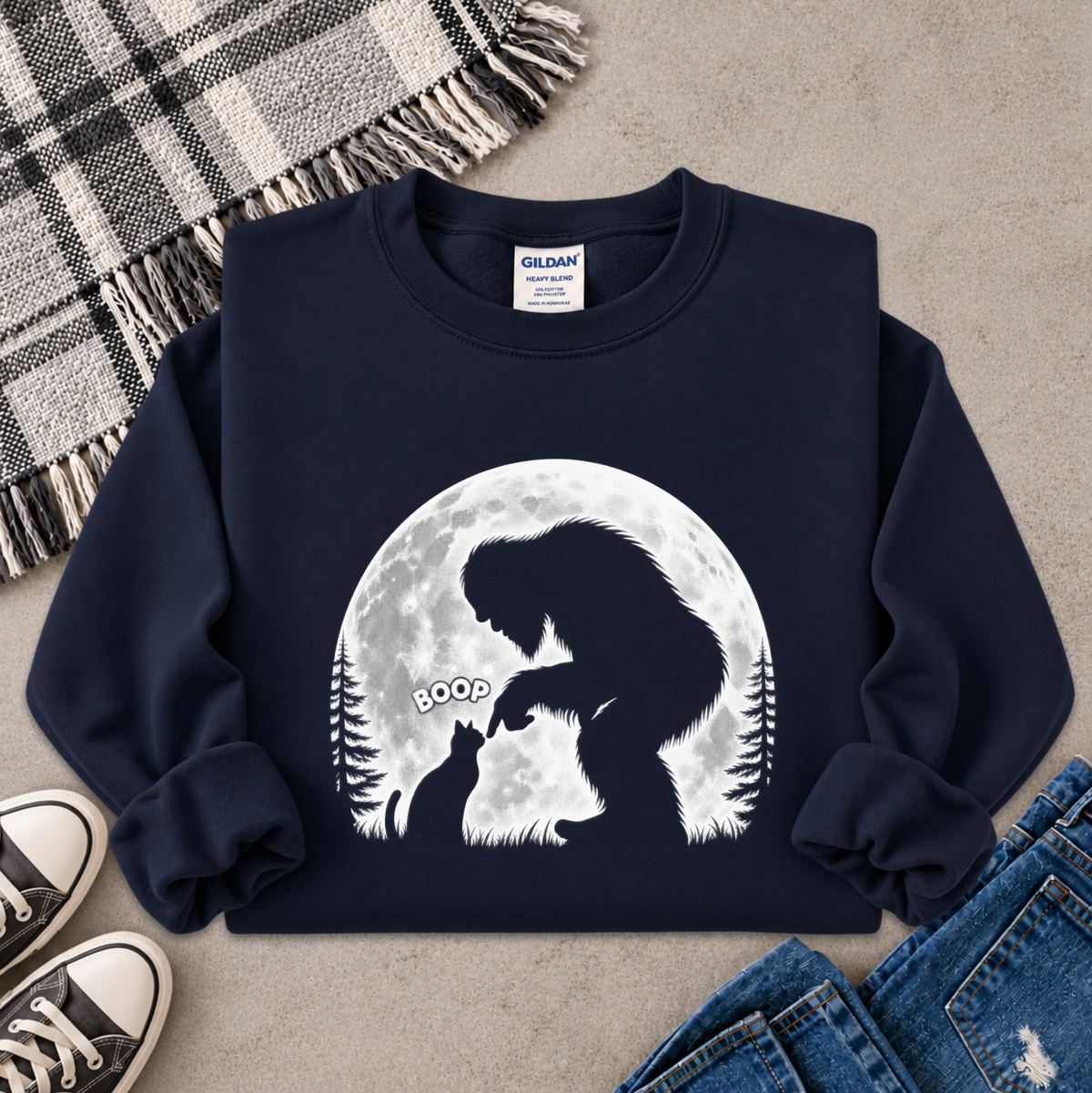 A navy blue crewneck sweatshirt with a white graphic of a Bigfoot silhouette looking at a cat in front of a full moon, styled in a flat lay with jeans, sneakers, and a plaid blanket on a concrete floor.