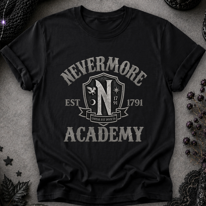 A black short-sleeve graphic T-shirt laid flat on a textured gray surface with dark decorative props around it. The shirt features a large distressed white graphic reading “NEVERMORE ACADEMY” with a crest-style “N” and “EST.