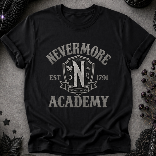 A black short-sleeve graphic T-shirt laid flat on a textured gray surface with dark decorative props around it. The shirt features a large distressed white graphic reading “NEVERMORE ACADEMY” with a crest-style “N” and “EST.