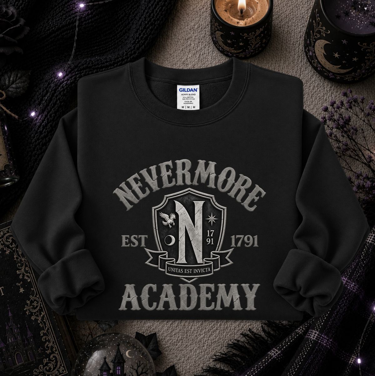 A black long-sleeve crewneck sweatshirt with a distressed gray graphic reading 'NEVERMORE ACADEMY' and a crest-style emblem, styled flat on a textured surface with candles, knit fabric, and gothic props around it.