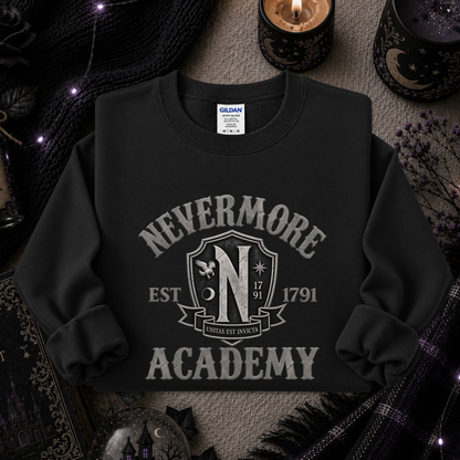 A black long-sleeve crewneck sweatshirt with a distressed gray graphic reading 'NEVERMORE ACADEMY' and a crest-style emblem, styled flat on a textured surface with candles, knit fabric, and gothic props around it.