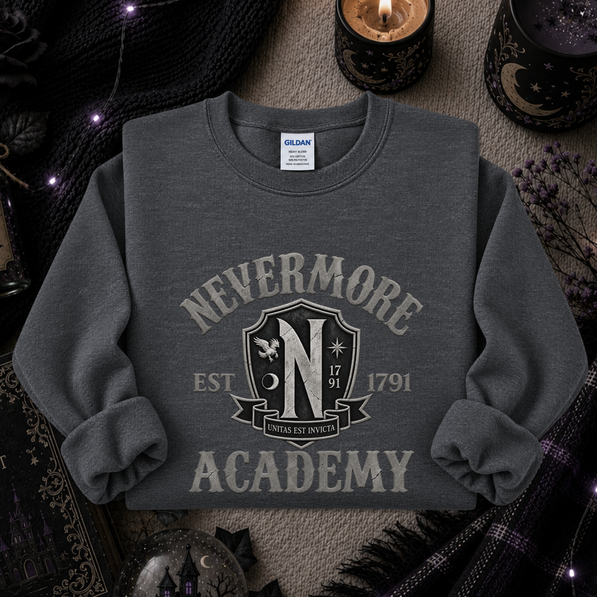 A dark gray crewneck sweatshirt featuring a distressed cream graphic that reads 'NEVERMORE ACADEMY' with a crest and 'EST 1791'. The sweatshirt is styled in a cozy, mystical flat-lay setting with candles, a black knit sweater, and dried flowers.