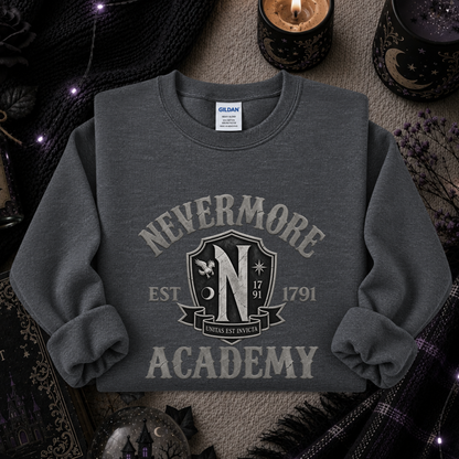 A dark gray crewneck sweatshirt featuring a distressed cream graphic that reads 'NEVERMORE ACADEMY' with a crest and 'EST 1791'. The sweatshirt is styled in a cozy, mystical flat-lay setting with candles, a black knit sweater, and dried flowers.